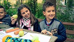 Kinder sitzen an einem Tisch und haben Hefte und frisches Gemüse vor sich.