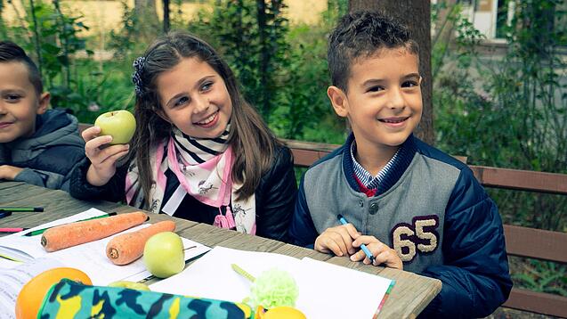 Kinder sitzen an einem Tisch und haben Hefte und frisches Gemüse vor sich.