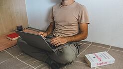 Ein junger Mann sitzt im Schneidersitz auf einem Bett und arbeitet am Laptop.