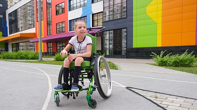 Ein Mädchen sitzt in einem Rollstuhl vor einem Schulgebäude.