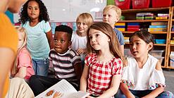 Eine Gruppe Grundschulkinder sitzt auf dem Boden, während ein Erwachsener etwas vorliest.