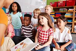 Eine Gruppe Grundschulkinder sitzt auf dem Boden, während ein Erwachsener etwas vorliest.