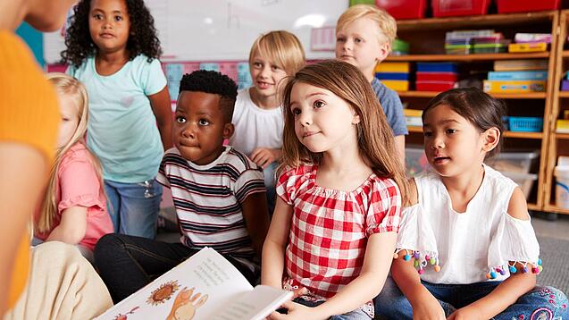 Eine Gruppe Grundschulkinder sitzt auf dem Boden, während ein Erwachsener etwas vorliest.
