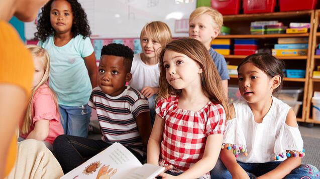 Eine Gruppe Grundschulkinder sitzt auf dem Boden, während ein Erwachsener etwas vorliest.