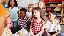 Eine Gruppe Grundschulkinder sitzt auf dem Boden, während ein Erwachsener etwas vorliest.
