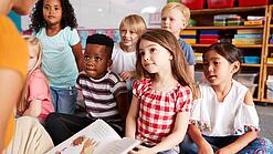 Eine Gruppe Grundschulkinder sitzt auf dem Boden, während ein Erwachsener etwas vorliest.