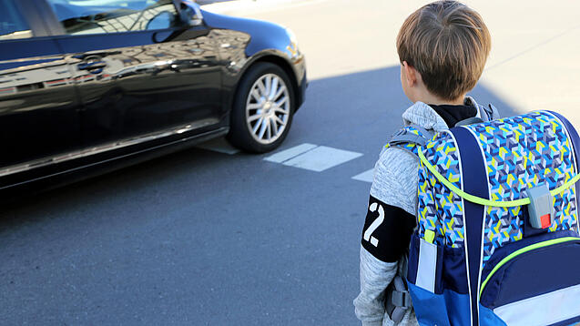 Ein junger Schüler steht mit seinem Schulranzen auf dem Rücken an einer Straße, auf der ein Auto fährt.