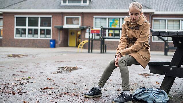 Ein junges Mädchen sitzt auf einer Bank auf dem Schulhof und schaut betreten zu Boden.