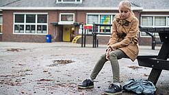 Ein junges Mädchen sitzt auf einer Bank auf dem Schulhof und schaut betreten zu Boden.