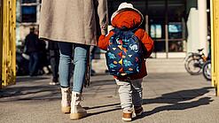 Ein junges Kind mit großem Rucksack geht an der Hand einer Erwachsenen.
