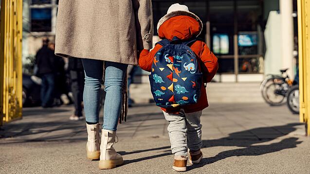 Ein junges Kind mit großem Rucksack geht an der Hand einer Erwachsenen.