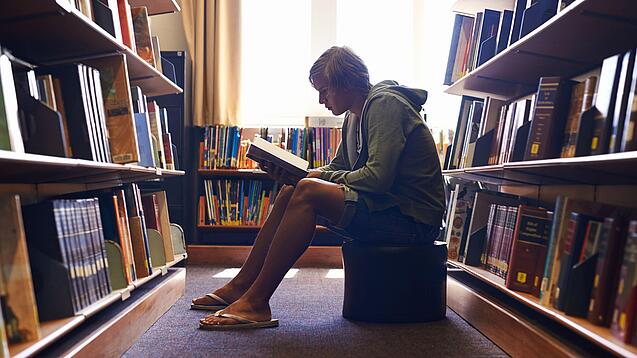 Ein Schüler sitzt auf einem Hocker zwischen Bücherregalen und liest ein Buch.