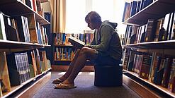 Ein Schüler sitzt auf einem Hocker zwischen Bücherregalen und liest ein Buch.