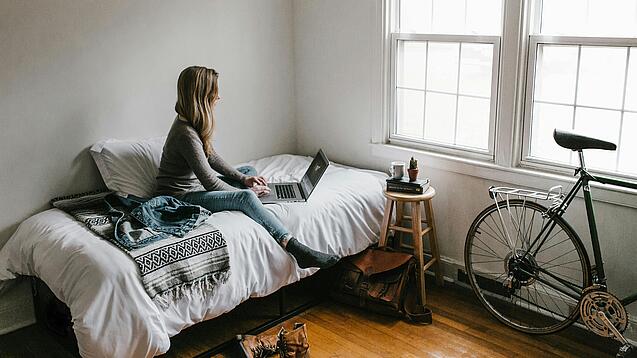Eine junge Frau sitzt auf einem Bett und arbeitet an ihrem Laptop, neben ihr ist ein Fahrrad an die Wand gelehnt.