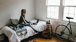Eine junge Frau sitzt auf einem Bett und arbeitet an ihrem Laptop, neben ihr ist ein Fahrrad an die Wand gelehnt.