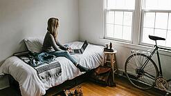 Eine junge Frau sitzt auf einem Bett und arbeitet an ihrem Laptop, neben ihr ist ein Fahrrad an die Wand gelehnt.