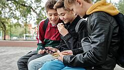 Drei Jungen sitzen auf einer Bank und schauen auf ein Smartphone.