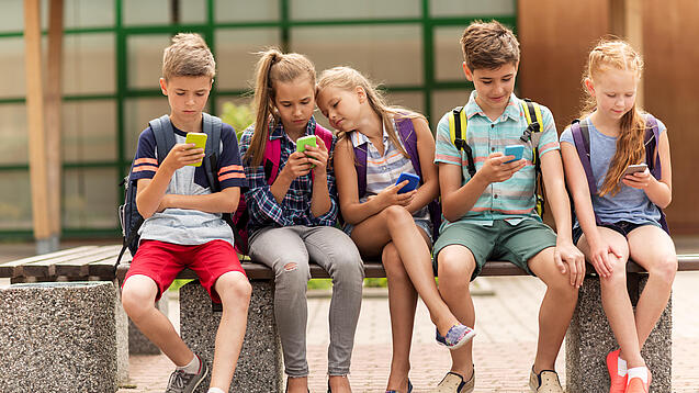 Kinder sitzen auf einer Bank auf dem Schulhof und schauen auf ihre Handys.