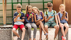 Kinder sitzen auf einer Bank auf dem Schulhof und schauen auf ihre Handys.