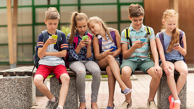 Kinder sitzen auf einer Bank auf dem Schulhof und schauen auf ihre Handys.