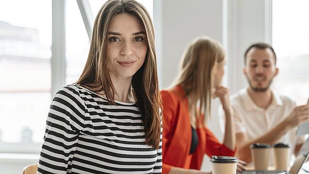 Eine junge Frau sitzt an einem Tisch und lächelt in die Kamera, im Hintergrund unterhalten sich zwei weitere Menschen.