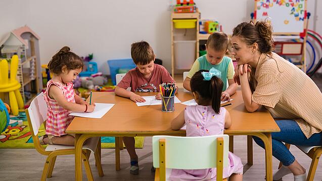 Kinder sitzen mit einer Erzieherin an einem Tisch und malen.