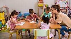 Kinder sitzen mit einer Erzieherin an einem Tisch und malen.