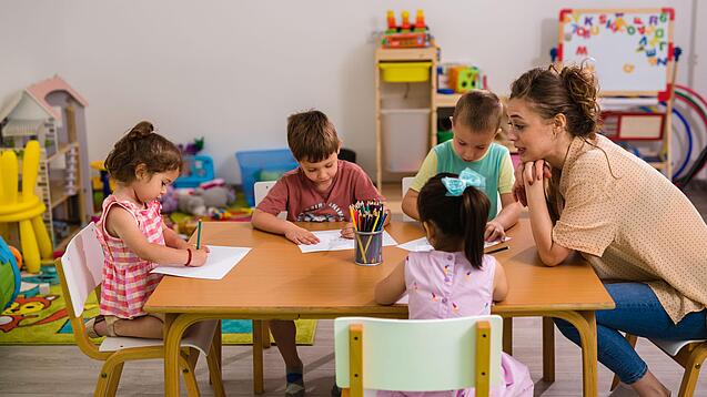 Kinder sitzen mit einer Erzieherin an einem Tisch und malen.