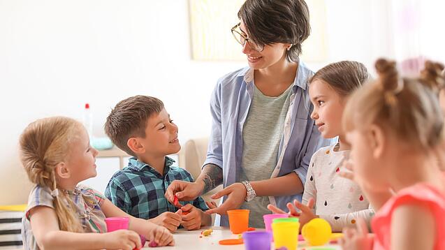 Eine Lehrerin ist mit Grundschulkindern um einen Tisch versammelt und sie bearbeiten etwas.