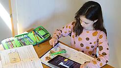 Ein junges Mädchen sitzt über ein Heft gebeugt an einem Tisch, in einer Hand hält sie ein Tablet, auf dem der Taschenrechner geöffnet ist.