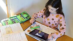 Ein junges Mädchen sitzt über ein Heft gebeugt an einem Tisch, in einer Hand hält sie ein Tablet, auf dem der Taschenrechner geöffnet ist.