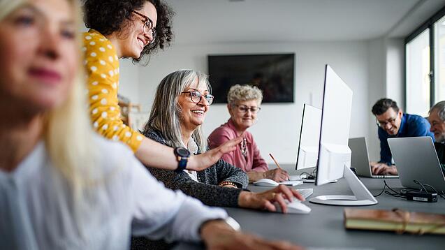 Ältere Personen sitzen an Computern. Eine Lehrerin erklärt ihnen etwas.