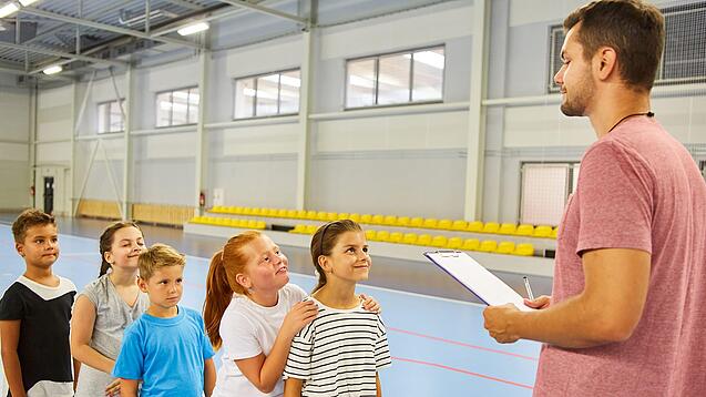 Ein junger Lehrer steht in einer Sporthalle vor einer Reihe Kinder im Grundschulalter.