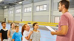 Ein junger Lehrer steht in einer Sporthalle vor einer Reihe Kinder im Grundschulalter.