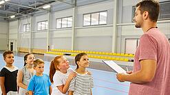 Ein junger Lehrer steht in einer Sporthalle vor einer Reihe Kinder im Grundschulalter.