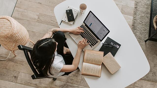 Eine junge Frau sitzt an einem Schreibtisch und arbeitet am Laptop, um sie herum liegen Bücher, ein Kalender, ein Handy und eine Tasse.