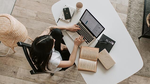 Eine junge Frau sitzt an einem Schreibtisch und arbeitet am Laptop, um sie herum liegen Bücher, ein Kalender, ein Handy und eine Tasse.