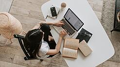 Eine junge Frau sitzt an einem Schreibtisch und arbeitet am Laptop, um sie herum liegen Bücher, ein Kalender, ein Handy und eine Tasse.