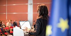 Eine Frau spricht an einem Podium. Rechts im Bild hängt eine Europa-Flagge.