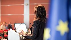 Eine Frau spricht an einem Podium. Rechts im Bild hängt eine Europa-Flagge.