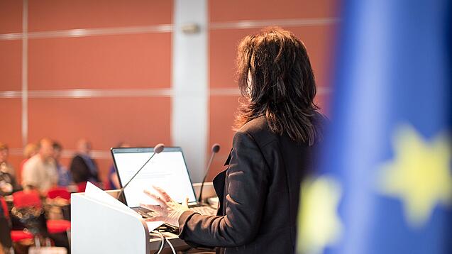 Eine Frau spricht an einem Podium. Rechts im Bild hängt eine Europa-Flagge.