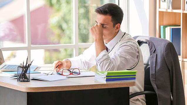 Ein junger Lehrer sitzt erschöpft am Schreibtisch, in der einen Hand hat er seine Brille, mit der anderen massiert er sich den Nasenrücken.