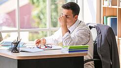 Ein junger Lehrer sitzt erschöpft am Schreibtisch, in der einen Hand hat er seine Brille, mit der anderen massiert er sich den Nasenrücken.