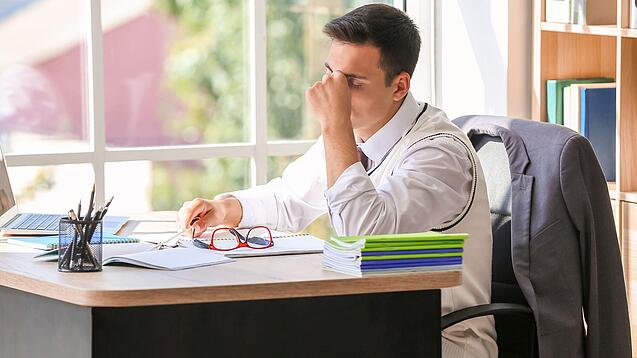 Ein junger Lehrer sitzt erschöpft am Schreibtisch, in der einen Hand hat er seine Brille, mit der anderen massiert er sich den Nasenrücken.