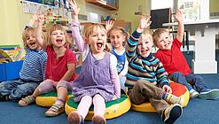 Gruppe Kinder sitzt auf Sitzkissen und hebt lachend die Hand.