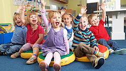 Gruppe Kinder sitzt auf Sitzkissen und hebt lachend die Hand.