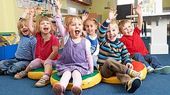 Gruppe Kinder sitzt auf Sitzkissen und hebt lachend die Hand.