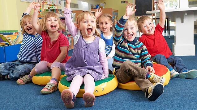 Gruppe Kinder sitzt auf Sitzkissen und hebt lachend die Hand.