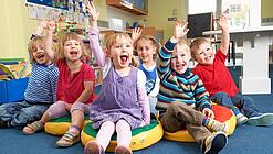 Gruppe Kinder sitzt auf Sitzkissen und hebt lachend die Hand.