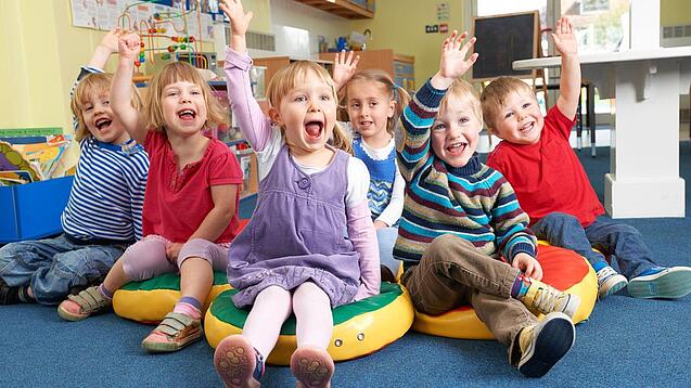 Gruppe Kinder sitzt auf Sitzkissen und hebt lachend die Hand.